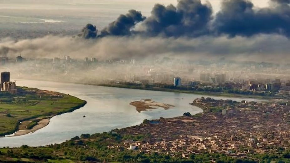 De gros nuages de fumée noire s'élèvent au-dessus d'une ville située sur un fleuve.