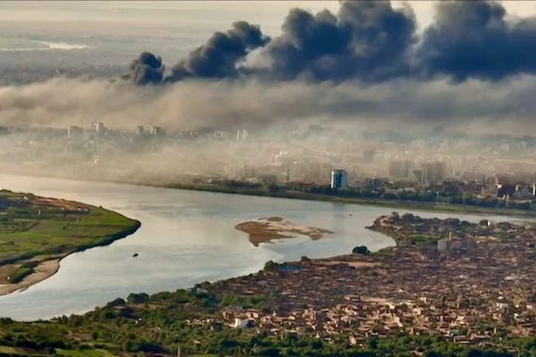 De gros nuages de fumée noire s'élèvent au-dessus d'une ville située sur un fleuve.