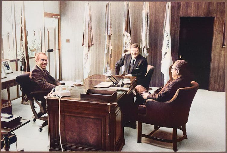 Henry Kissinger, Richard Nixon and John Wayne sit around a desk in an office in front of flags.