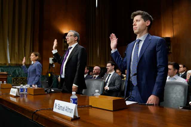 two men and a woman in business suits stand behind a long desk with their right hands raised