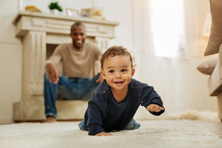 A smiling baby crawling towards the camera in the foreground, and a young man smiling in the background