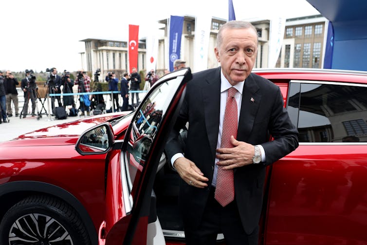 A man in suit and tie exits a red car.