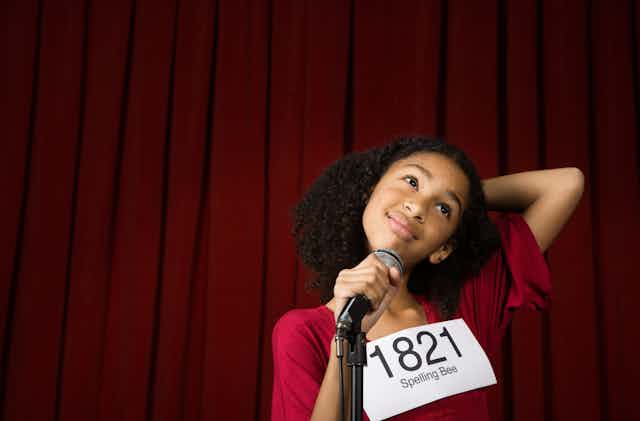 A young girl with a spelling bee contestant sign with the number 1821 holds a microphone on stage with one hand and has her other hand behind her head as she appears to be thinking about how to spell a word.