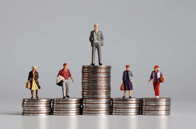 Tiny human figures standing on coins: one man on a tall stack, and several people on shorter stacks