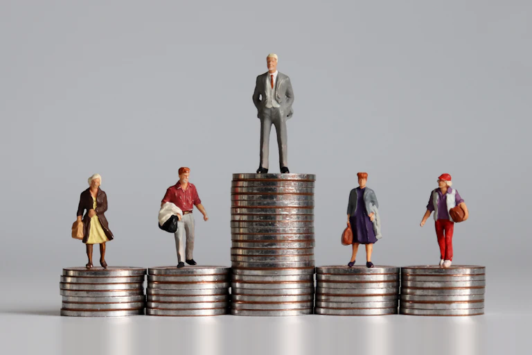 Tiny human figures standing on coins: one man on a tall stack, and several people on shorter stacks