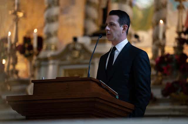Kendall Roy (Jeremy Strong) stands behind a lectern in a black suit. 