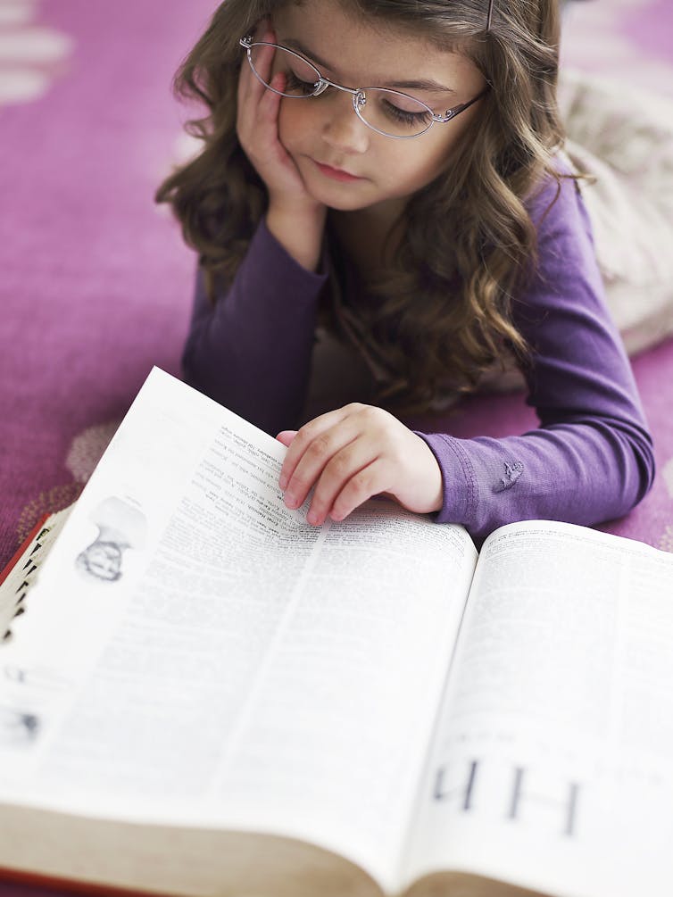 A girl reads a dictionary as she lies on the floor.