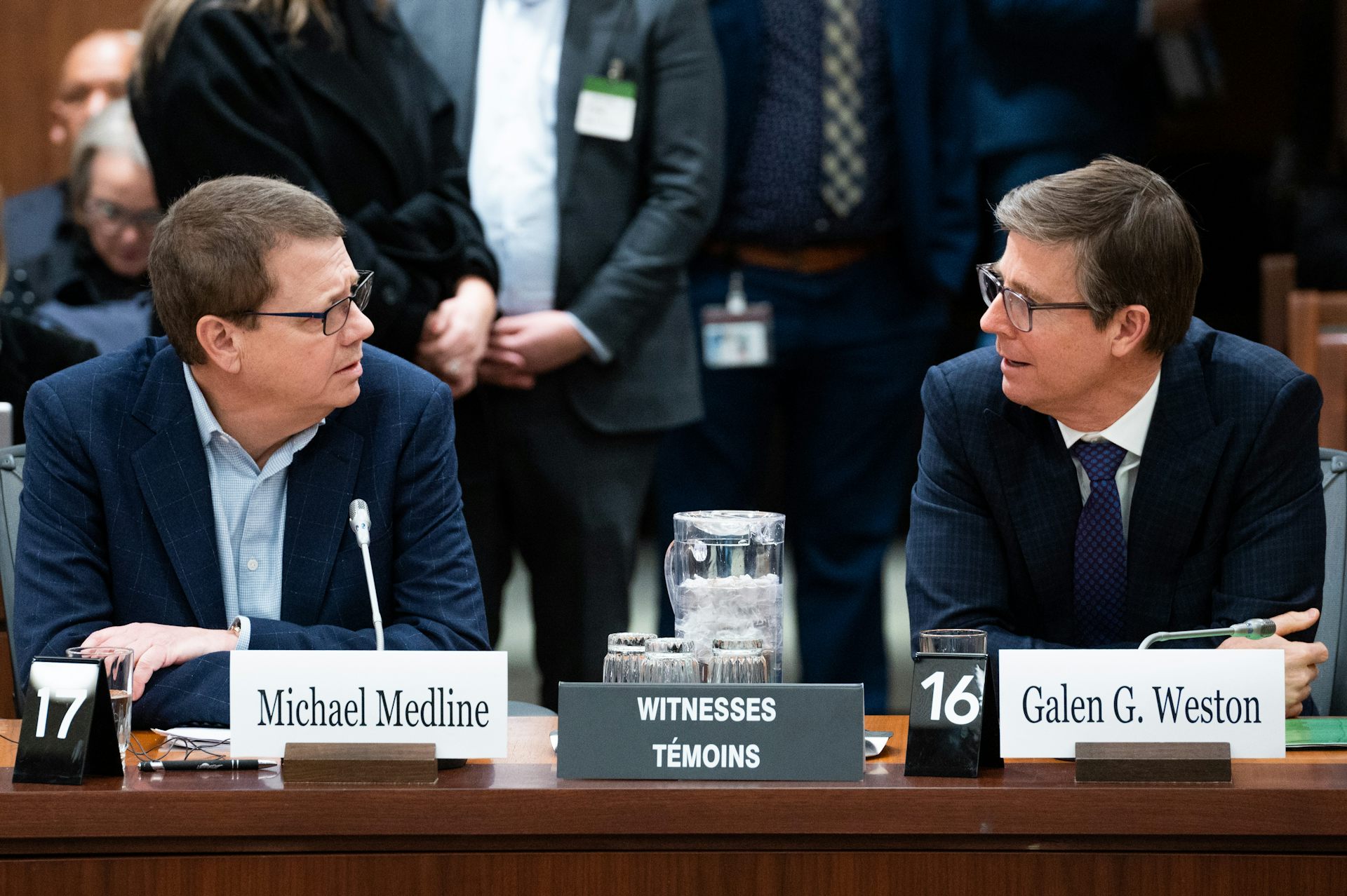 Two middle-aged white men wearing glasses and navy suits talk to one another while seated from behind a desk.