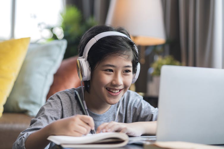 A middle school-aged boy with headphones looks at a laptop screen. He is smiling and writing in a notebook.