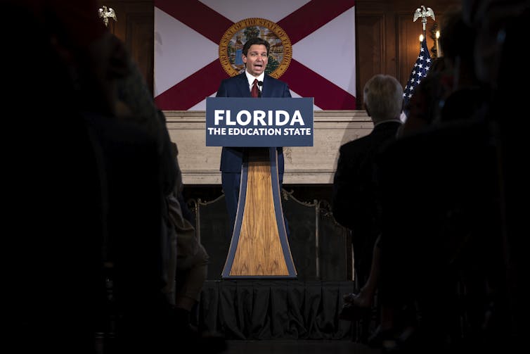A man in a dark suit standing at a lectern with the sign 'Florida, the education state' on it.