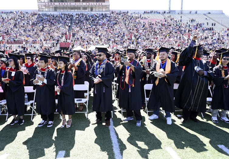 Muchos graduados con batas académicas pasan junto a una gran multitud en un estadio.