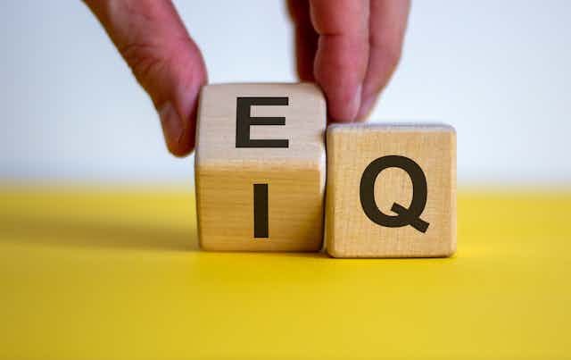 A hand rolling a small wooden cube with the letters I and E on it so that the E will be more prominently displayed. A cube with the letter Q on it sits to its right.
