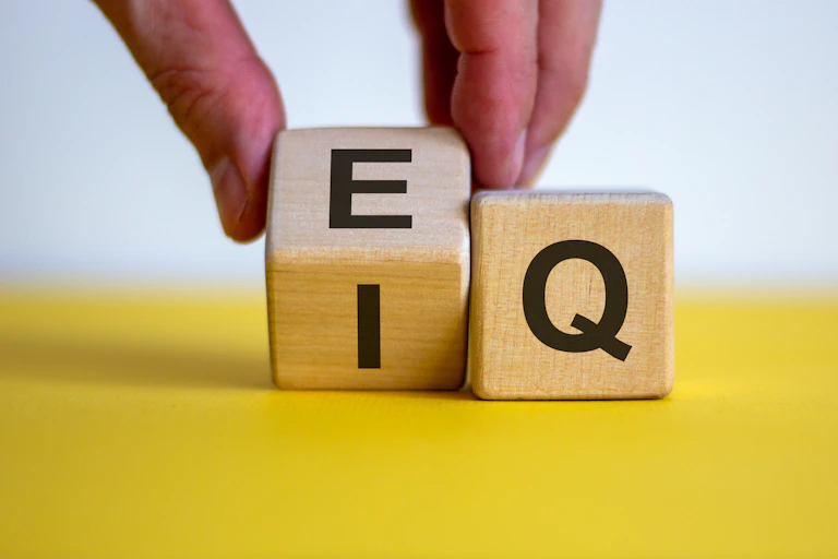 A hand rolling a small wooden cube with the letters I and E on it so that the E will be more prominently displayed. A cube with the letter Q on it sits to its right.