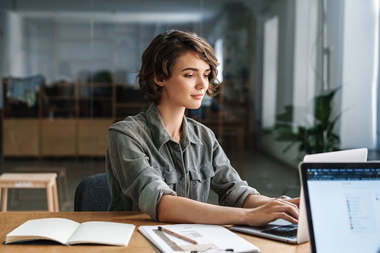 A young woman looking content as she types on an open laptop at a desk in an office.