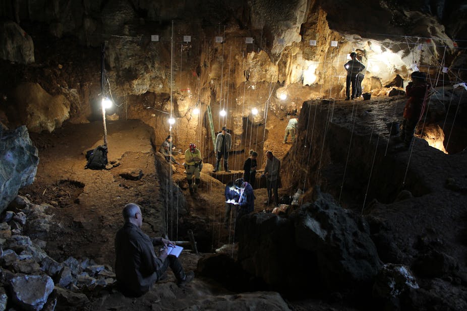 A photo shows several archaeologists at work in a cave hung with lights.