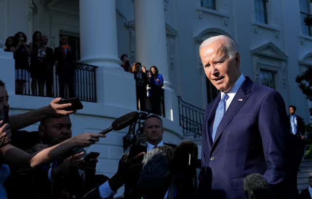 an elderly white man in a blue suit looks ahead as microphones are thrust in his face in front of a white ornate building