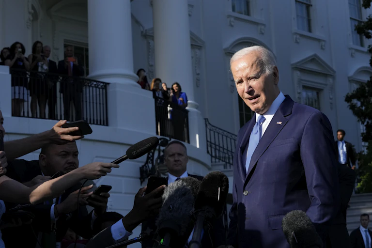 an elderly white man in a blue suit looks ahead as microphones are thrust in his face in front of a white ornate building