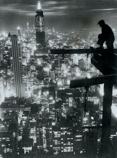 Man sits on construction scaffolding overlooking New York City skyline.