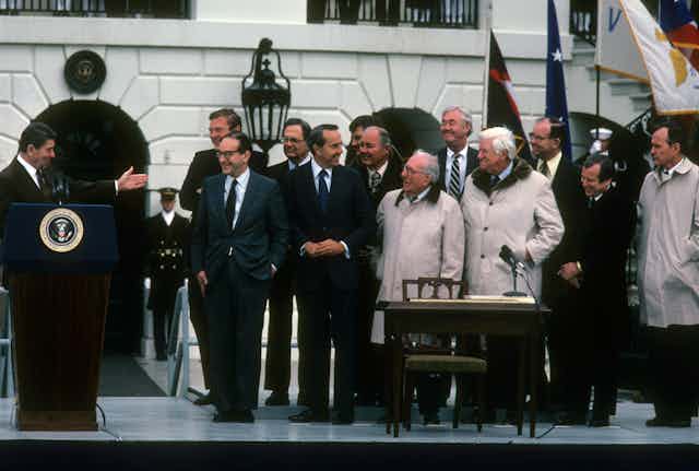 President Reagan, standing behind a lectern outside a building, gestures to gathered dignitaries.