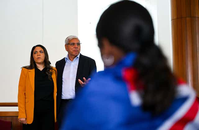 Jacinta Price and Warren Mundine stand speaking in front of a young First Nations person who wears an Australian flag around their shoulders.