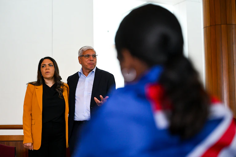 Jacinta Price and Warren Mundine stand speaking in front of a young First Nations person who wears an Australian flag around their shoulders.