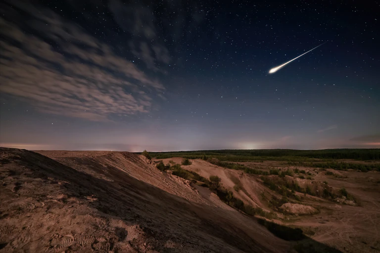 A vast, desert landscape stretches towards a dark blue horizon. A blast of light is heading for the ground.