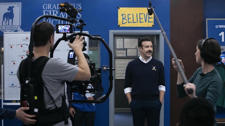 Un actor ante cámaras y micrófonos rodando una escena de una serie ante la puerta de un despacho que tiene un cartel colocado con la palabra 'cree' en inglés.