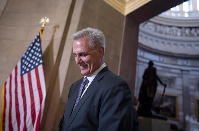 a white man wearing a suit smiles looking down next to an American flag and a statue in the distance.