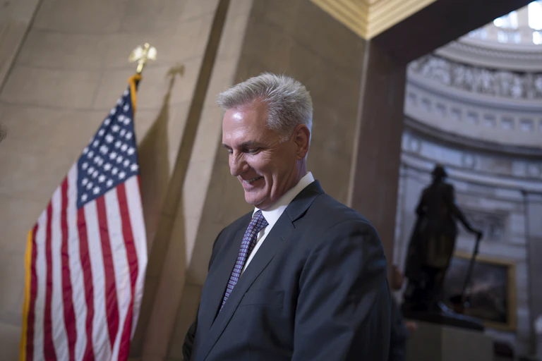 a white man wearing a suit smiles looking down next to an American flag and a statue in the distance.
