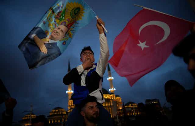 A man on friend's shoulders hold aloft a flag with a man's image on. Next to him is a Turkish flag.