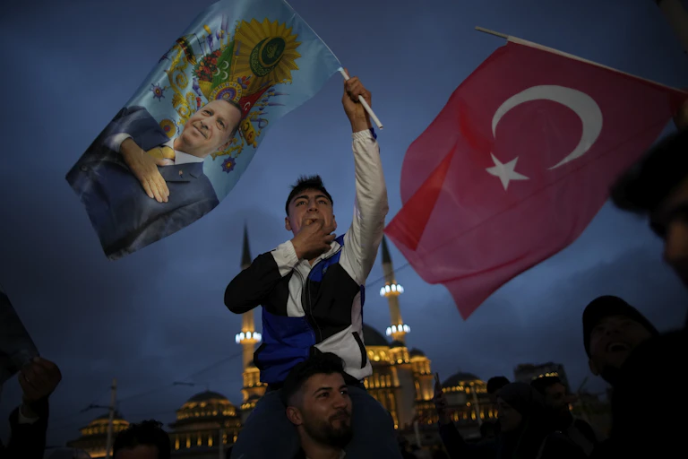 A man on friend's shoulders hold aloft a flag with a man's image on. Next to him is a Turkish flag.