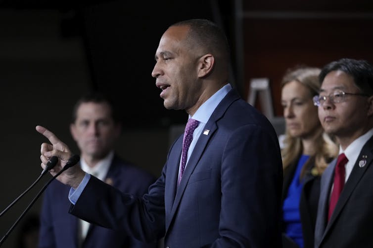 A black man in a suit speaks at a podium in front of several other people