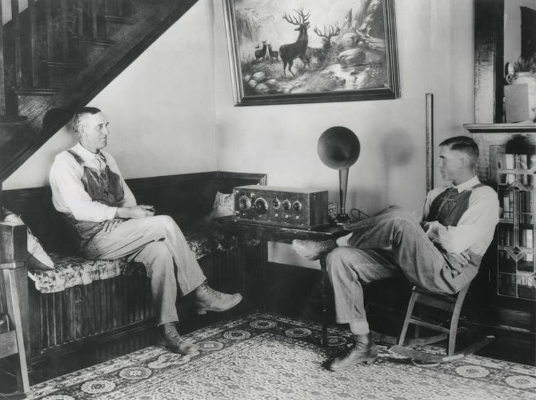 A black and white photo of two farmers sitting in a living room listening to a large wooden radio with a bullhorn attached.