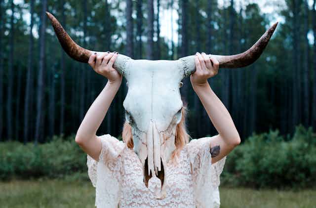 a woman in a lacy dress holding a goat skull over her face, against a forest backdrop