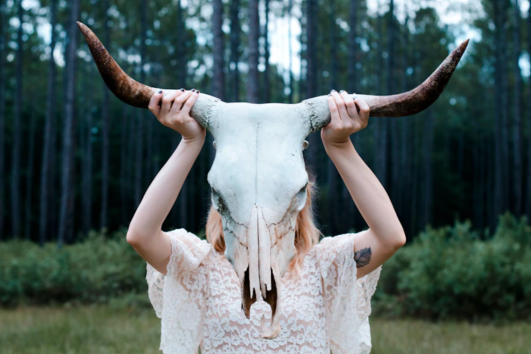 a woman in a lacy dress holding a goat skull over her face, against a forest backdrop