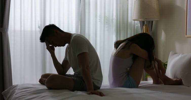 A man and a woman sitting a bed with their backs to each other and their heads bowed
