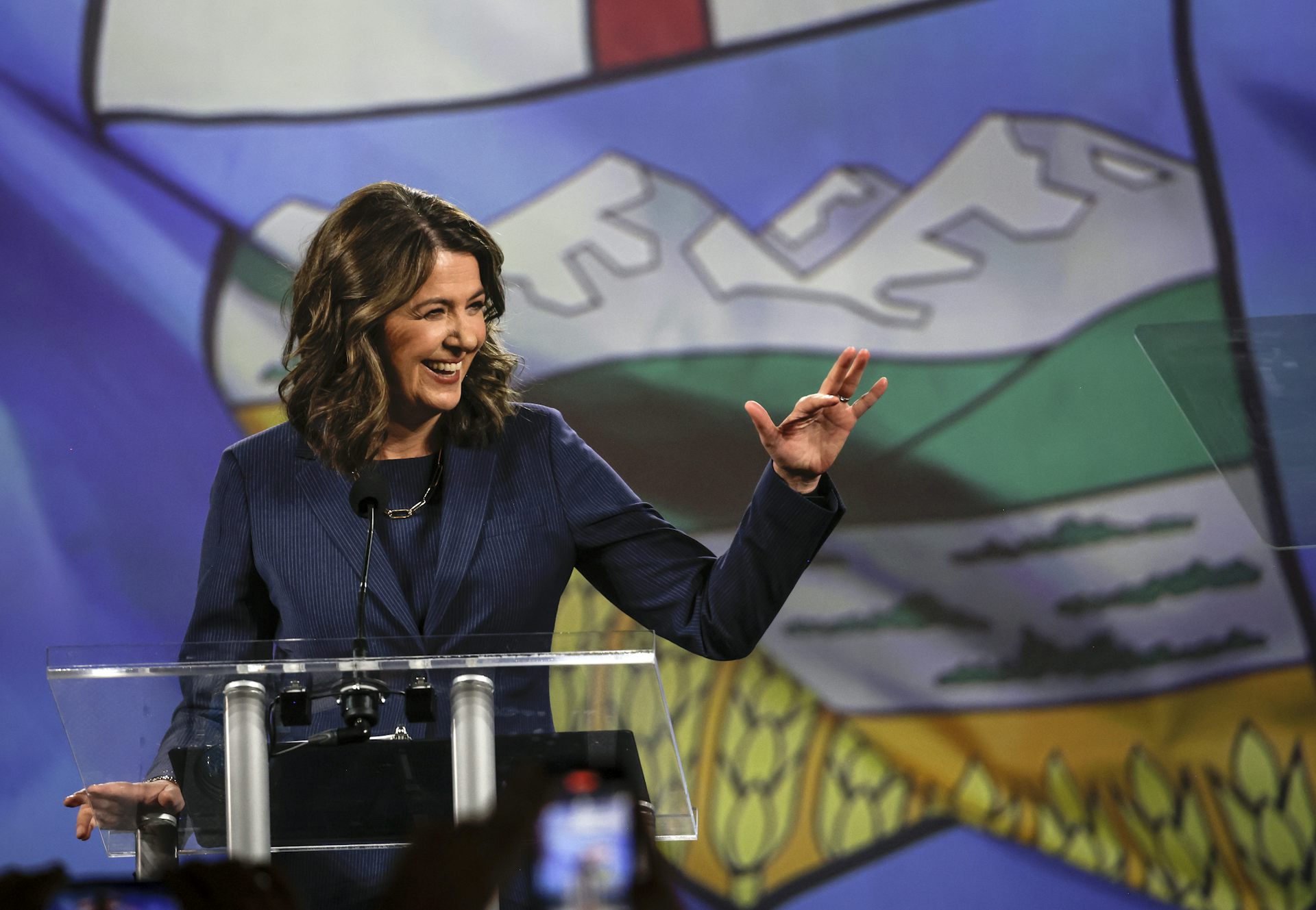 A dark-haired woman smiles and waves from behind a podium.