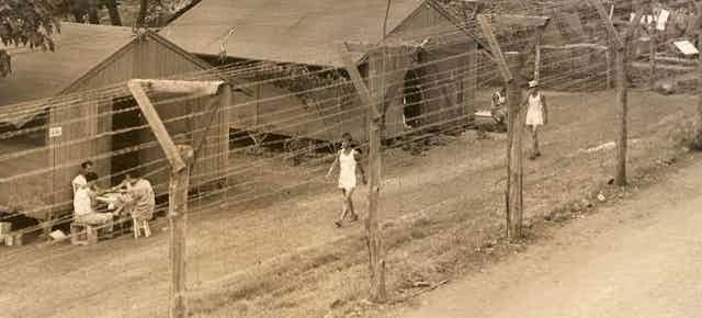 Several men are walking behind a barbed wire fence and wooden buildings.