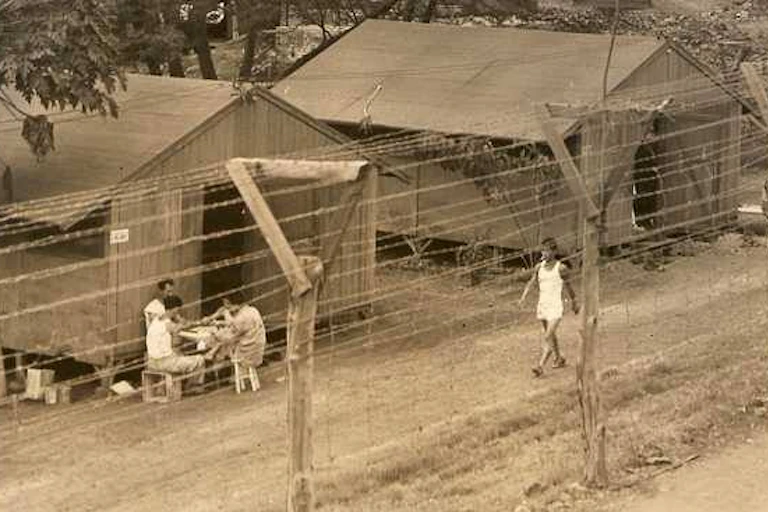 Several men are walking behind a barbed wire fence and wooden buildings.