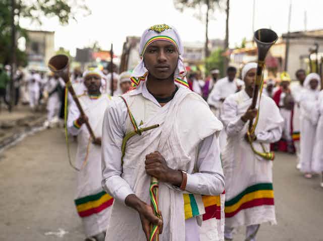 A group of men in traditional Ethiopian robes move in a procession down a street, holding musical instruments