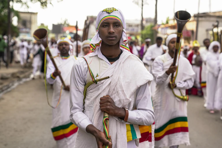A group of men in traditional Ethiopian robes move in a procession down a street, holding musical instruments