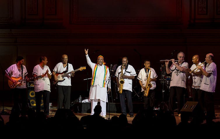 A band on stage with a man in Ethiopian robes in the centre, an arm raised and a smile on his face.