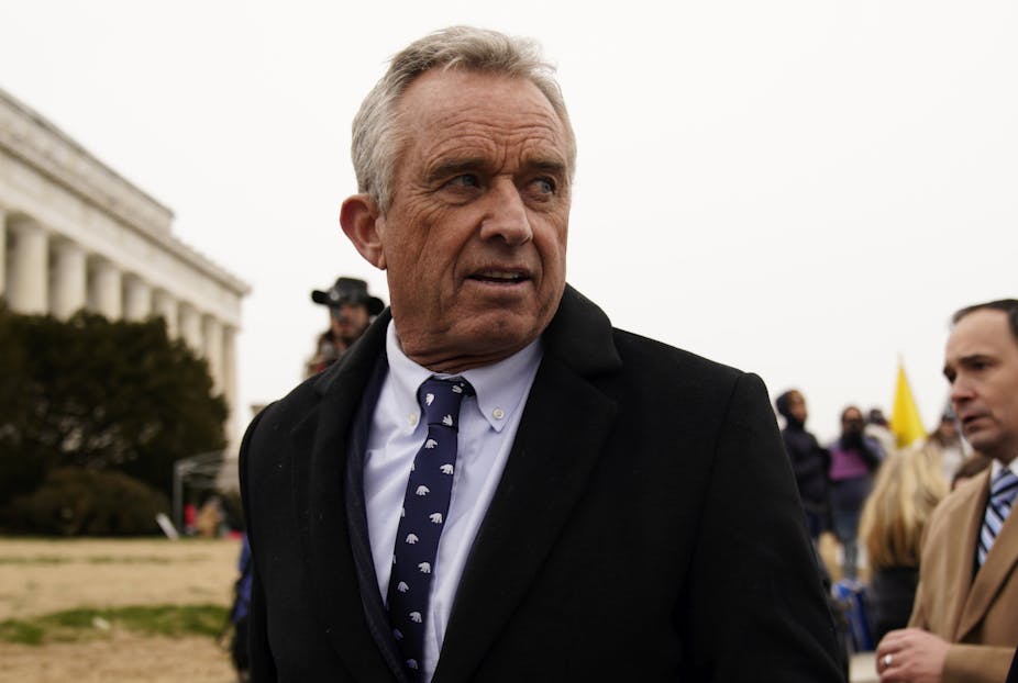 Photo of RFK Jr wearing a suit and tie and looking over his shoulder, there are other marchers near him and the columns of the Lincoln Memorial are visible in the background