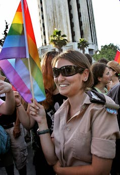 A woman in sunglasses and a tan military uniform smiles and holds a rainbow-striped flag.