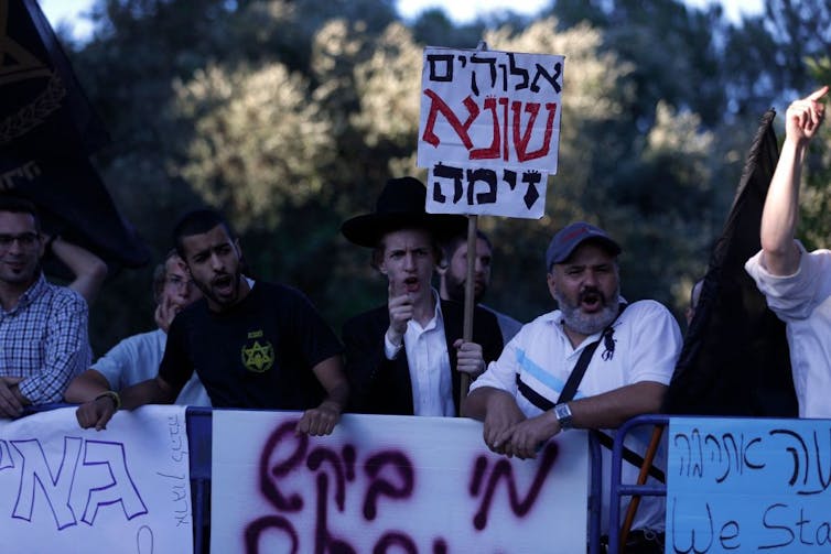 Angry-looking men holding signs in Hebrew shout during a protest.
