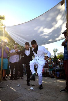 Two men embrace as they stomp drinking glasses on the ground. One wears a black suit and one wears a white suit.