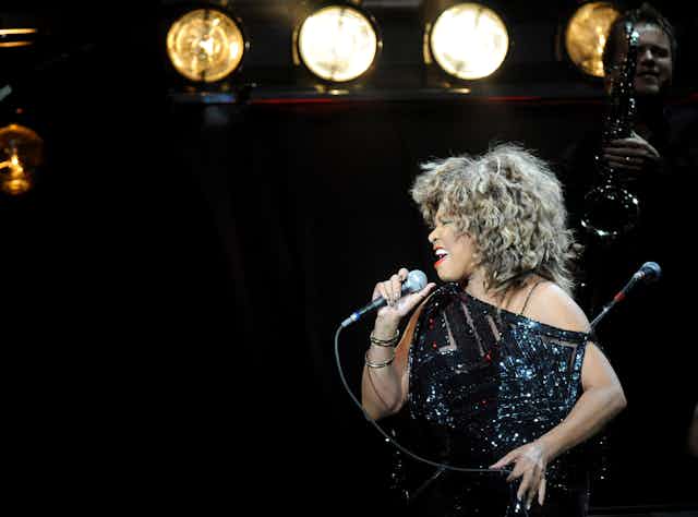Tina Turner with curly hair and black sequin dress singing into a microphone.