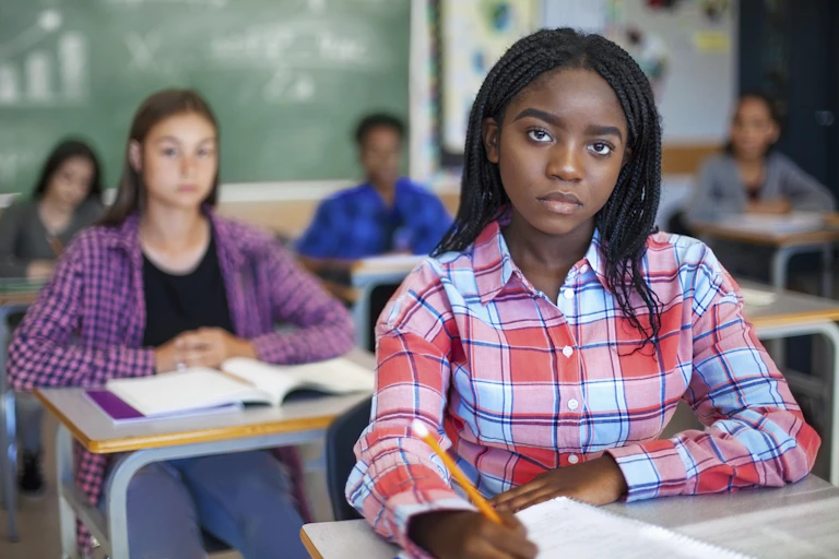 A Black female student wearing light blue and light red plaid shirt looks forward. Several students and a chalkboard are in the background.