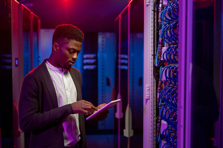 A young man in a trendy but professional outfit taps on an ipad while standing next to an open cabinet of wires and cables. the hallway is cast in red and blue light.