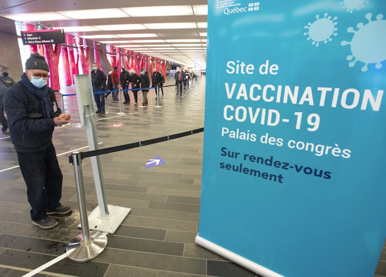 A lineup of people in face masks behind a sign for a COVID-19 vaccination clinic.